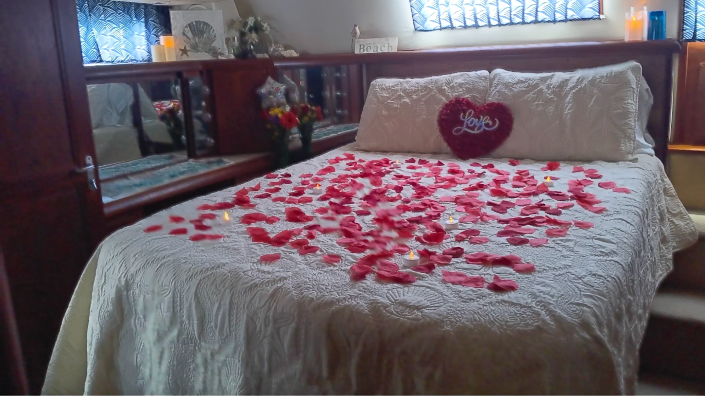 Bed with rose petals and a heart-shaped pillow with 'love' in a cozy, decorated room.
