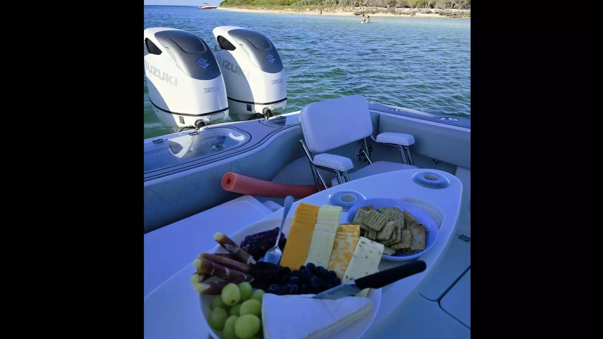 Center console boat with twin Suzuki outboards, stern seating and charcuterie spread anchored near a tropical shoreline.