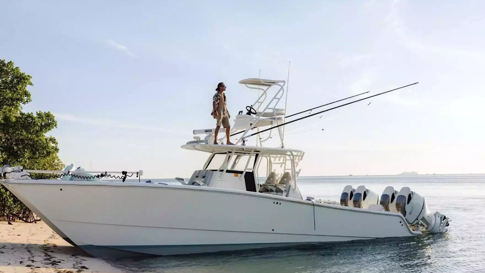 Center console boat with tower, fishing rods and multiple outboard engines positioned along a tropical shoreline.
