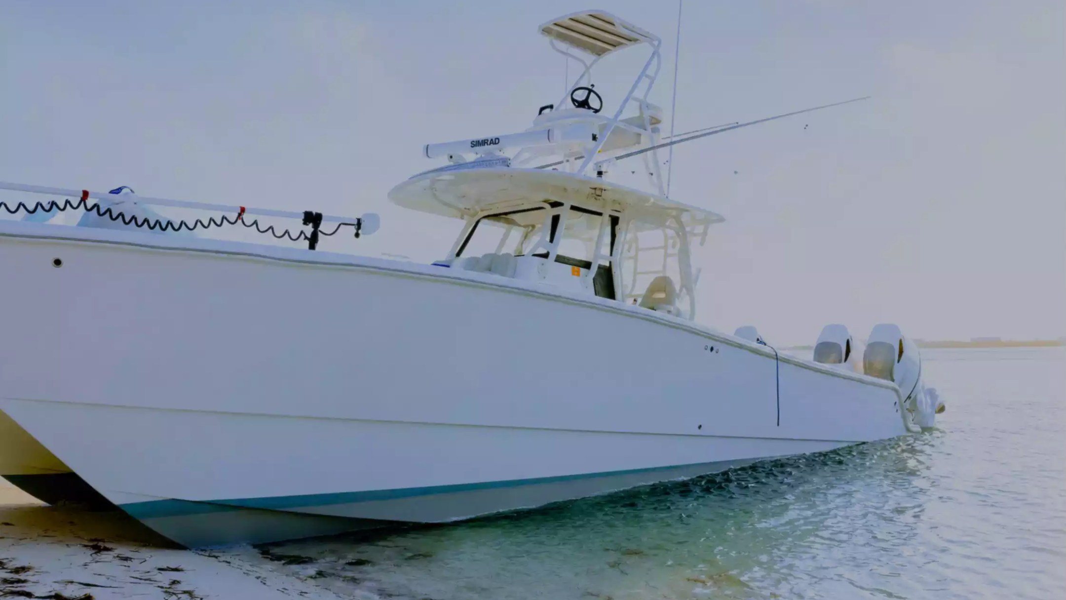 Center console fishing boat with elevated tower, fishing rods and multiple outboard engines beached along a quiet shoreline.