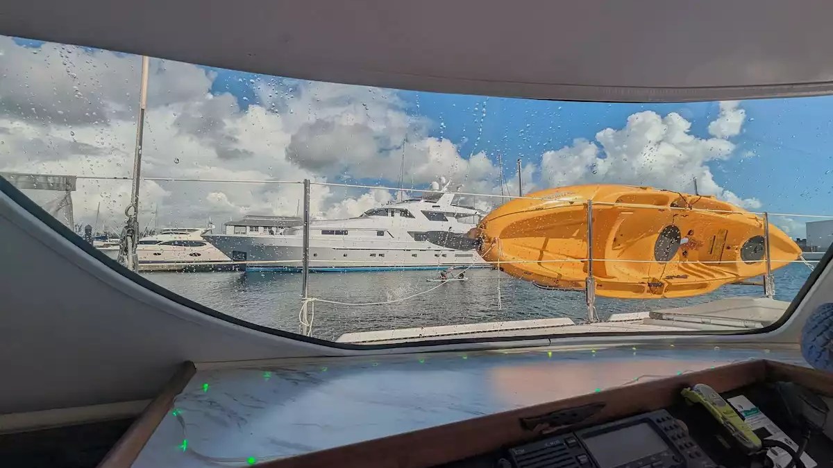 Sailing catamaran panoramic salon window overlooking an orange kayak and marina with yachts under blue skies.